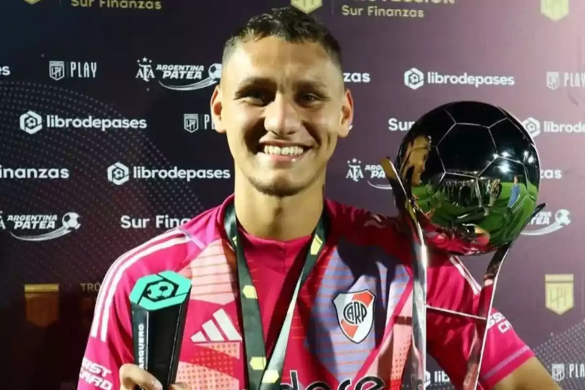 Portero de River Plate sonríe mientras sostiene un trofeo y un premio individual frente a un fondo con logotipos de patrocinadores. Portero de River Plate sonríe mientras sostiene un trofeo y un premio individual frente a un fondo con logotipos de patrocinadores.