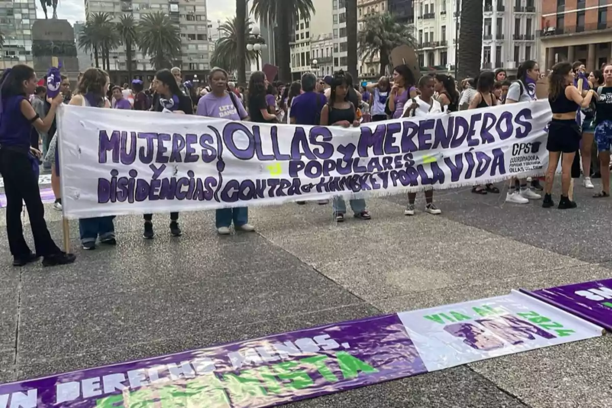 Group of women holding a banner at a demonstration that reads: Women, community kitchens, popular soup kitchens, dissidents against hunger and for life, in a square surrounded by buildings and palm trees. Group of women holding a banner at a demonstration that reads: Women, community kitchens, popular soup kitchens, dissidents against hunger and for life, in a square surrounded by buildings and palm trees.