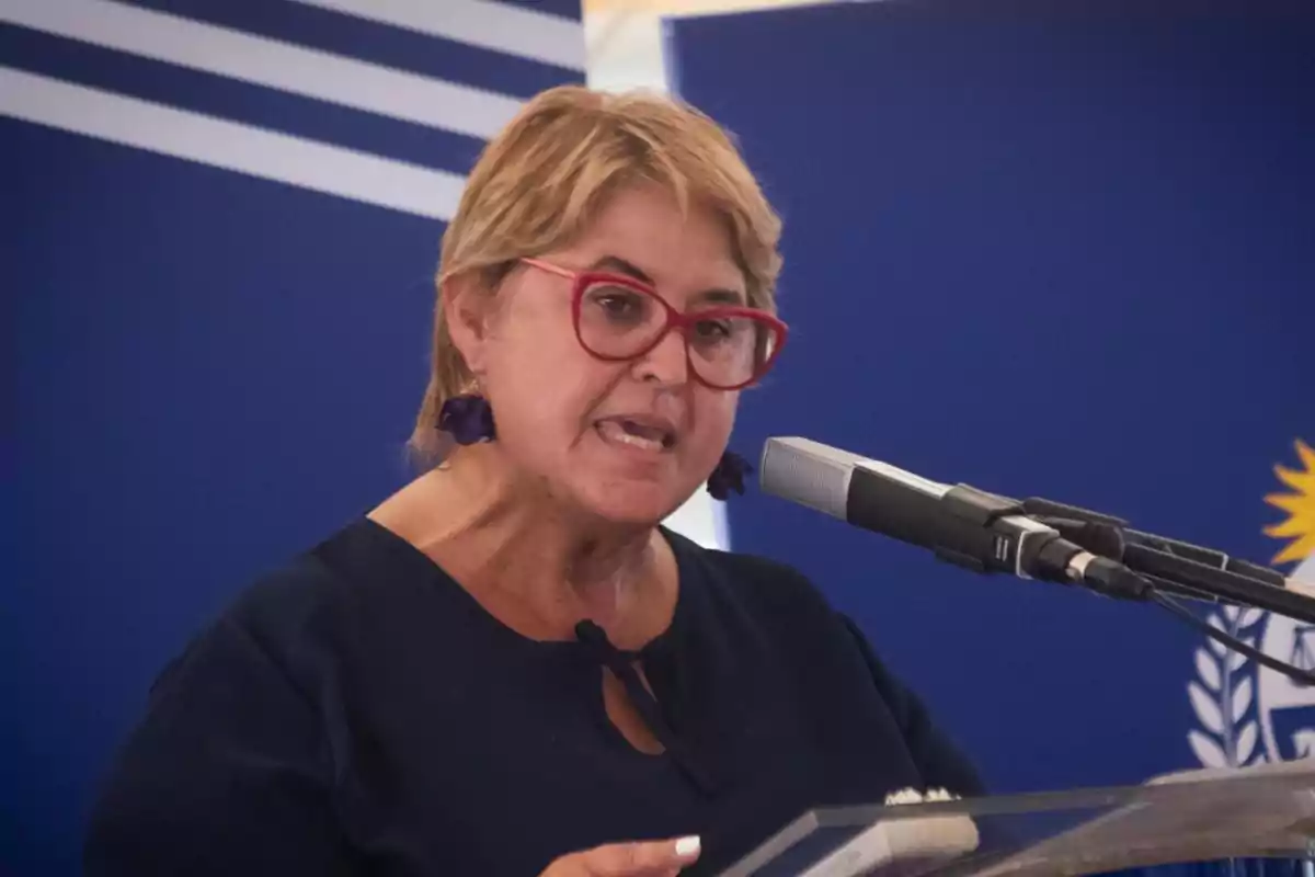 Mujer de cabello corto y gafas rojas hablando frente a un micrófono en un podio con fondo azul institucional Mujer de cabello corto y gafas rojas hablando frente a un micrófono en un podio con fondo azul institucional