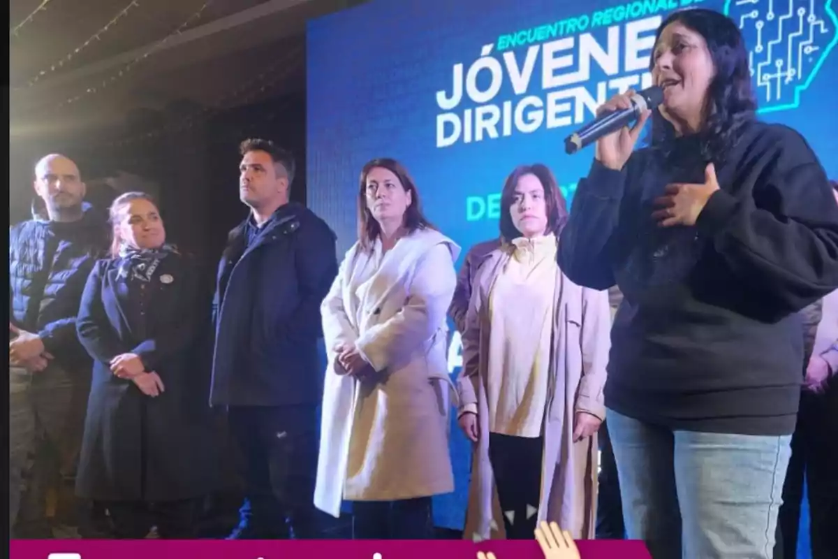 A group of people is standing on a stage while a woman in front speaks into a microphone, and in the background there is a screen with the text A group of people is standing on a stage while a woman in front speaks into a microphone, and in the background there is a screen with the text