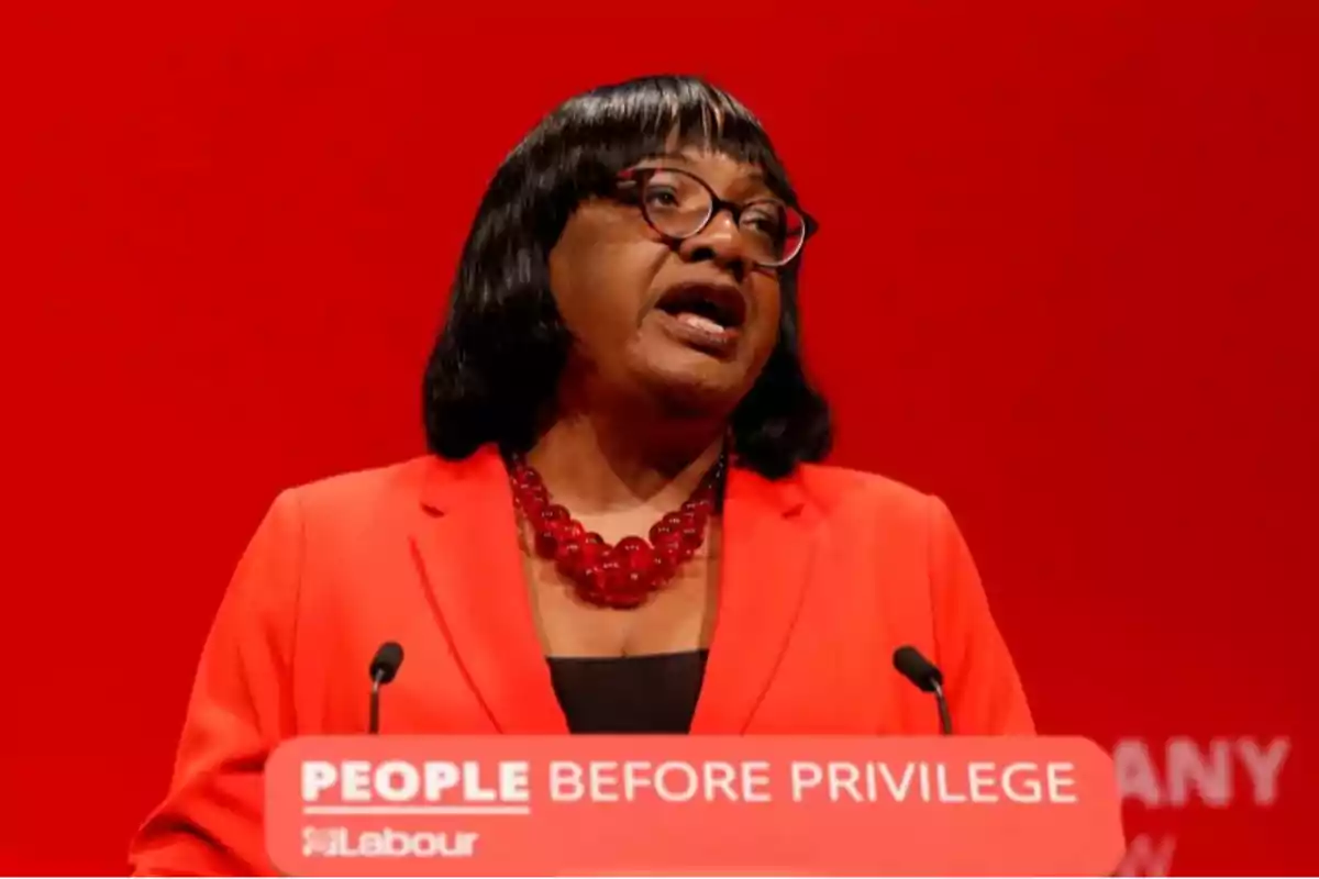 Woman with glasses and a red necklace speaking at a podium with a red background and a sign that says PEOPLE BEFORE PRIVILEGE