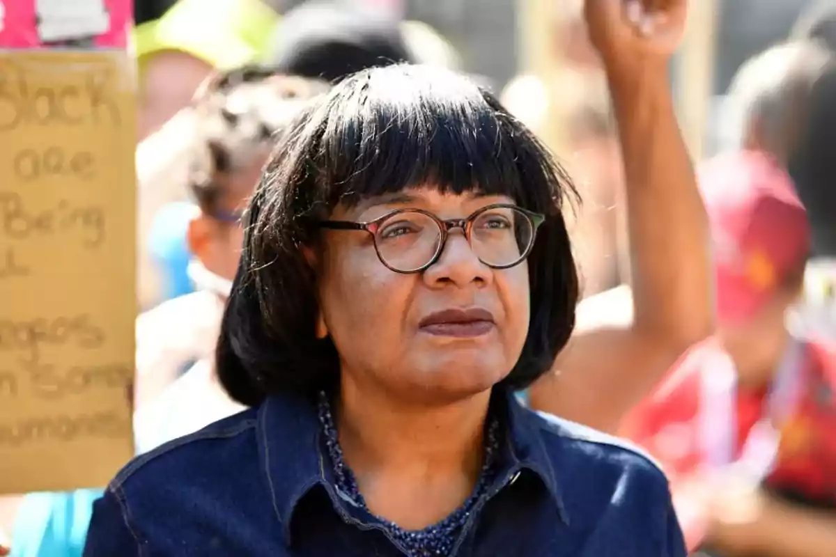 Mujer con gafas y cabello corto oscuro observa con expresión seria durante una manifestación rodeada de personas y un cartel al fondo Mujer con gafas y cabello corto oscuro observa con expresión seria durante una manifestación rodeada de personas y un cartel al fondo