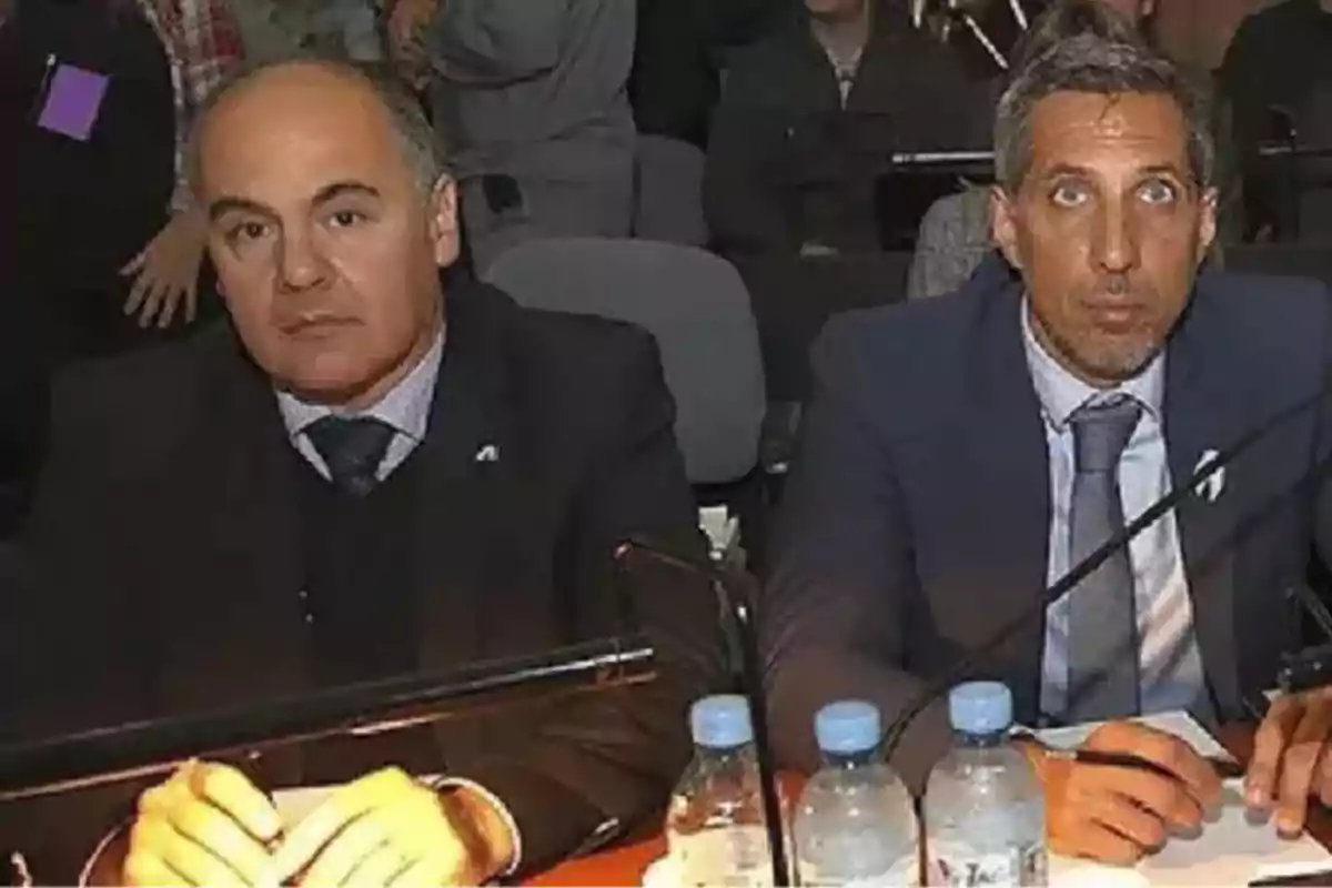 Dos hombres con traje y corbata están sentados en una mesa con botellas de agua y papeles, en lo que parece ser una reunión o conferencia, rodeados de otras personas al fondo. Dos hombres con traje y corbata están sentados en una mesa con botellas de agua y papeles, en lo que parece ser una reunión o conferencia, rodeados de otras personas al fondo.