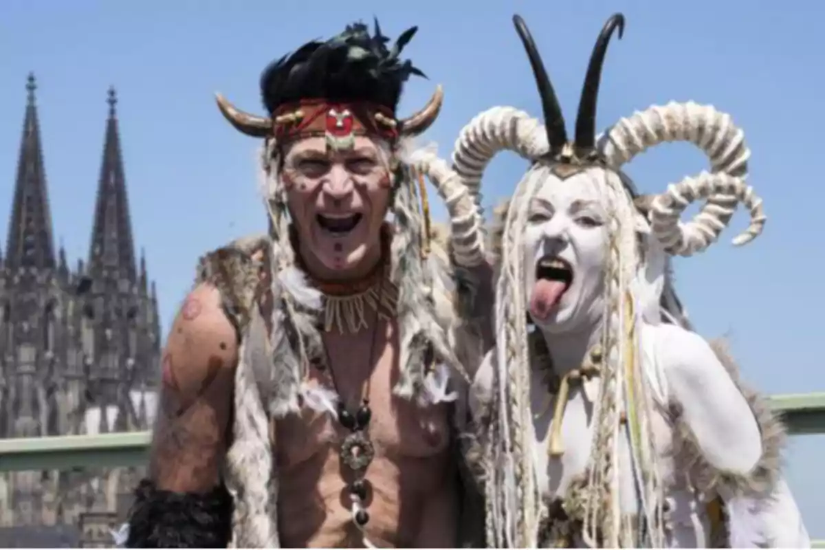 Two people dressed in tribal attire and horns pose in front of a Gothic cathedral under a clear sky. Two people dressed in tribal attire and horns pose in front of a Gothic cathedral under a clear sky.