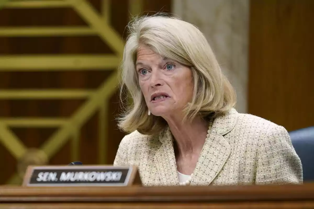 Mujer de cabello rubio y expresión seria sentada en una sala de reuniones con un letrero que dice Sen. Murkowski Mujer de cabello rubio y expresión seria sentada en una sala de reuniones con un letrero que dice Sen. Murkowski