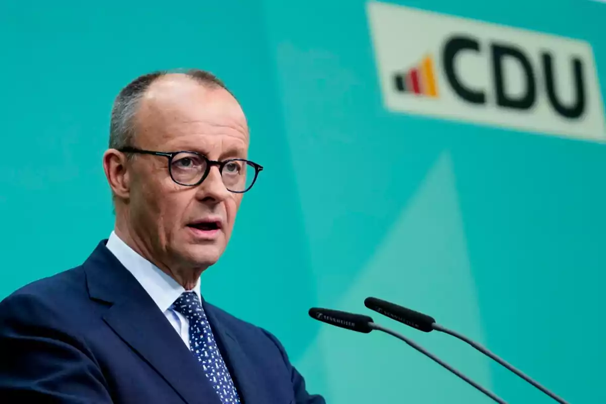 Man in a suit and glasses speaking in front of microphones with the CDU logo in the background on a turquoise wall Man in a suit and glasses speaking in front of microphones with the CDU logo in the background on a turquoise wall
