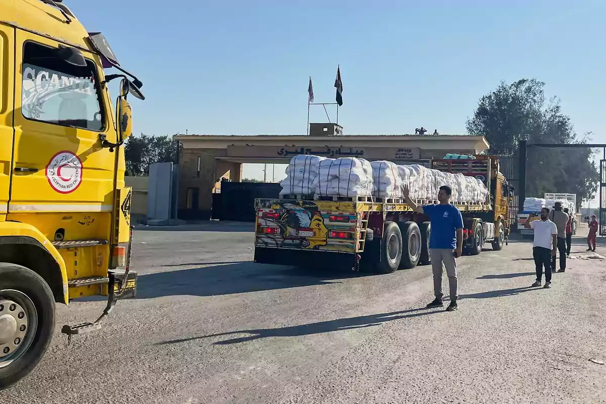Yellow trucks loaded with humanitarian aid move toward a border crossing as several people walk and oversee the operation under a clear sky.
