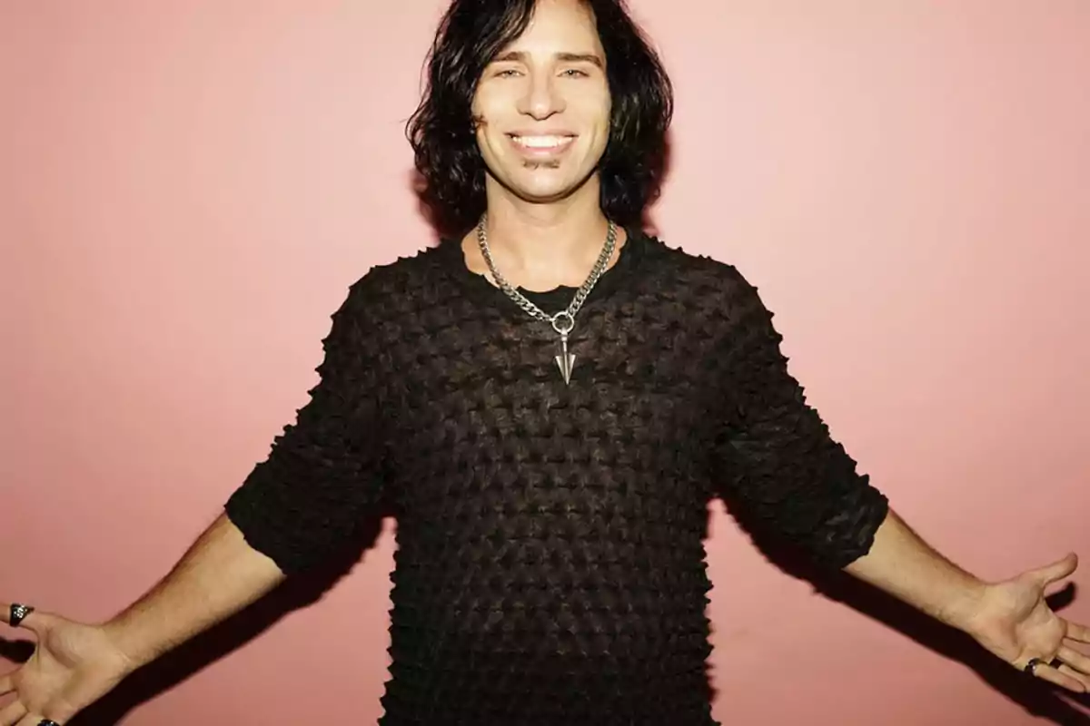 Hombre de cabello oscuro y ondulado sonríe con los brazos abiertos vistiendo una camiseta negra de textura peculiar y un collar plateado frente a un fondo rosa