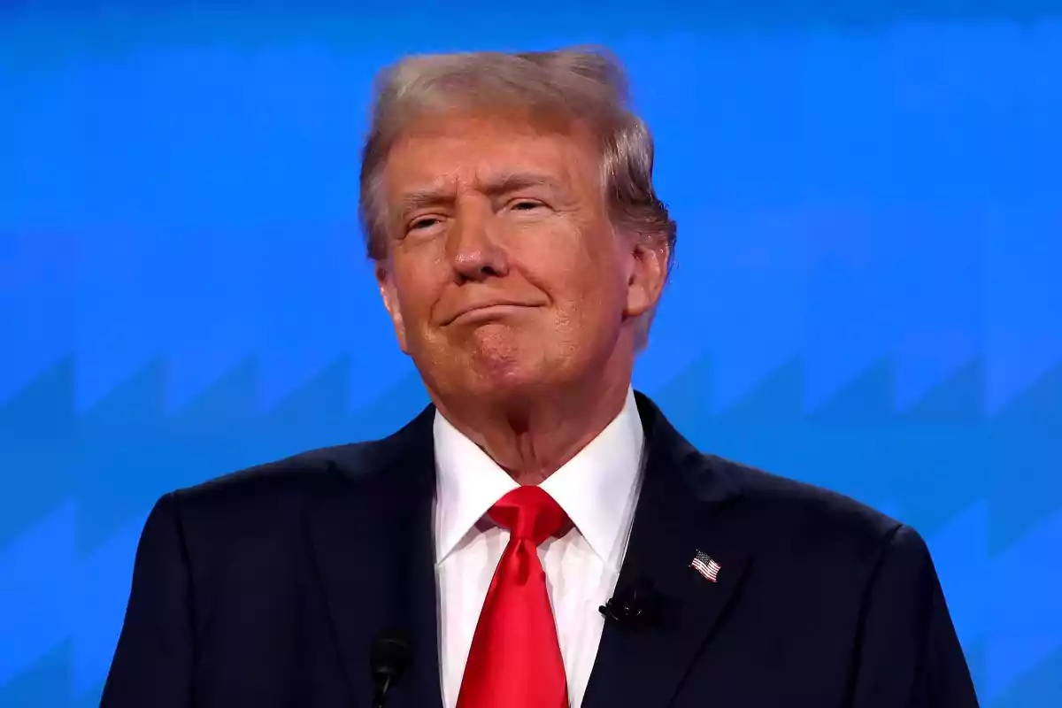 Blond-haired man in a dark suit, white jersey, and red tie, standing in front of a blue background