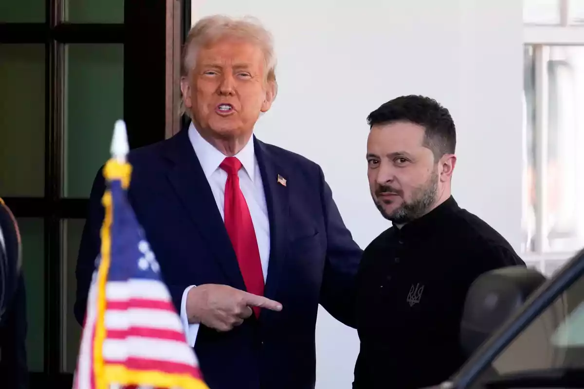 Dos hombres de pie juntos en una entrada, uno con traje azul y corbata roja señalando al otro, mientras una bandera de Estados Unidos aparece en primer plano.