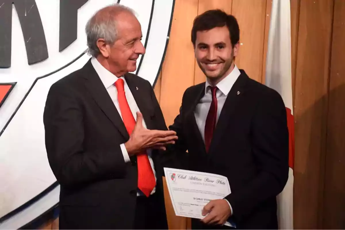 Dos hombres con traje sonríen mientras uno de ellos sostiene un diploma frente a un fondo con el escudo de un club deportivo. Dos hombres con traje sonríen mientras uno de ellos sostiene un diploma frente a un fondo con el escudo de un club deportivo.