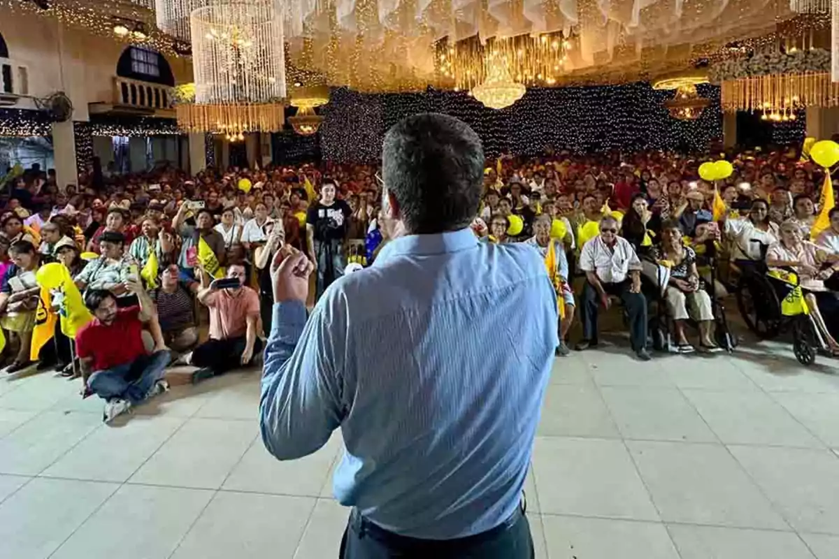 Persona de espaldas hablando frente a una multitud en un salón decorado con luces y candelabros mientras el público sostiene globos y banderas amarillas
