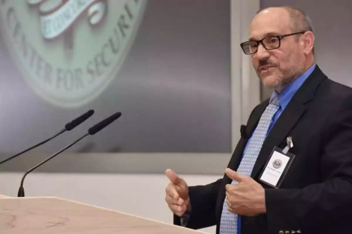 Hombre con gafas y traje hablando en un podio frente a dos micrófonos con un logo institucional de fondo Hombre con gafas y traje hablando en un podio frente a dos micrófonos con un logo institucional de fondo