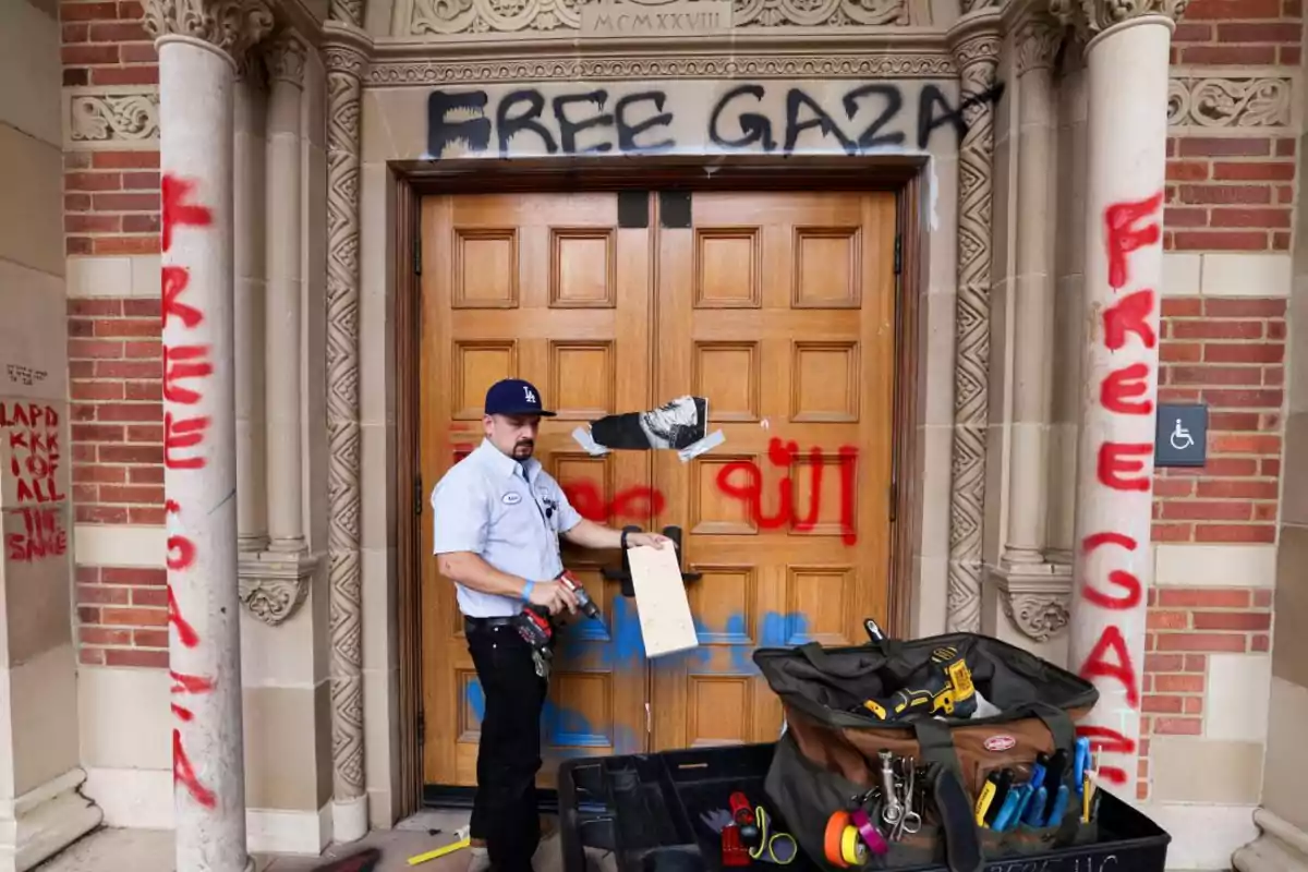 Un trabajador con herramientas repara la puerta de un edificio antiguo que ha sido vandalizado con grafitis de mensajes políticos en las paredes y columnas. Un trabajador con herramientas repara la puerta de un edificio antiguo que ha sido vandalizado con grafitis de mensajes políticos en las paredes y columnas.