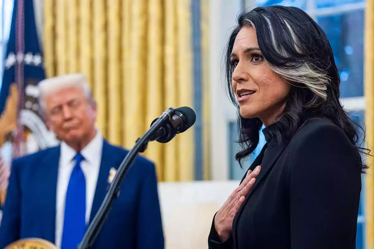 A dark-haired woman with a white streak is speaking into a microphone while a blond man in a blue suit watches her from the background in a room with golden curtains. A dark-haired woman with a white streak is speaking into a microphone while a blond man in a blue suit watches her from the background in a room with golden curtains.