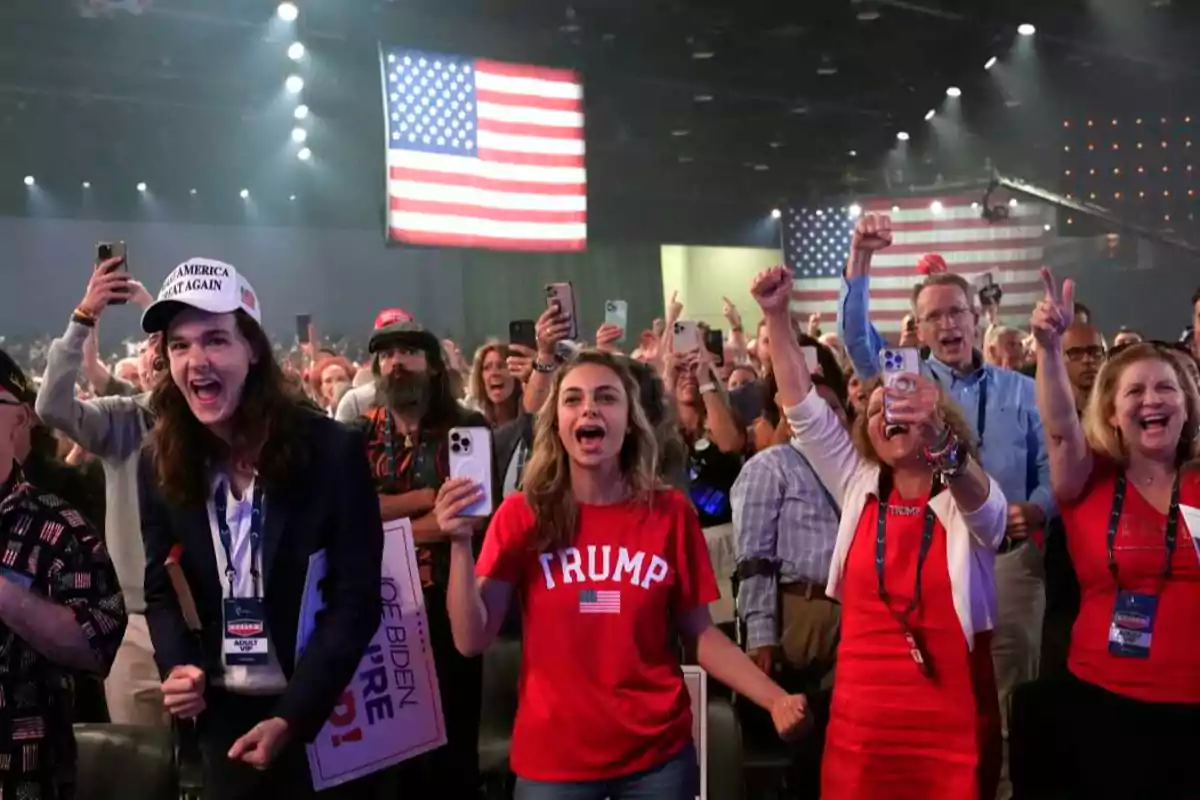 Grupo de personas animadas en un evento político con camisetas y gorras de apoyo a Trump, algunas levantando los brazos y tomando fotos, con banderas de Estados Unidos al fondo. Grupo de personas animadas en un evento político con camisetas y gorras de apoyo a Trump, algunas levantando los brazos y tomando fotos, con banderas de Estados Unidos al fondo.