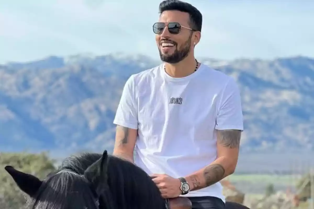 Man with a beard and sunglasses riding a black horse with mountains in the background and wearing a white t-jersey Man with a beard and sunglasses riding a black horse with mountains in the background and wearing a white t-jersey