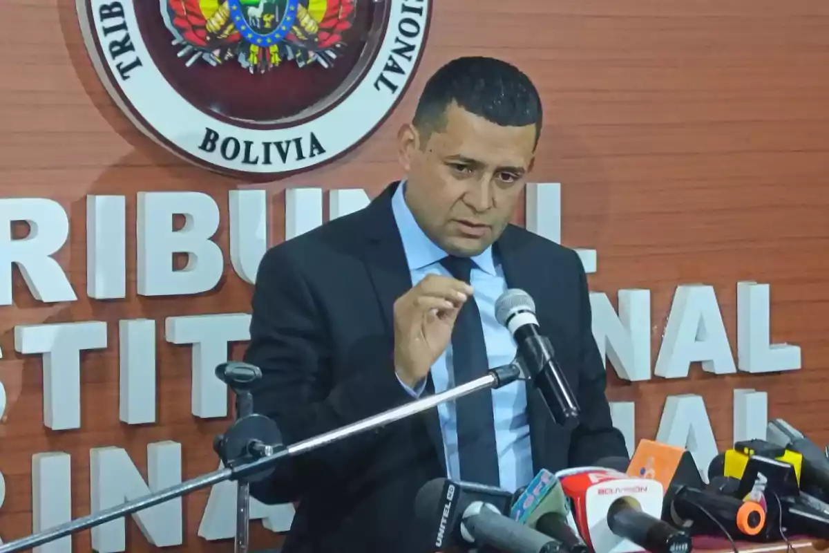 Un hombre de traje oscuro habla en una conferencia de prensa frente a varios micrófonos y un fondo con el escudo de Bolivia y letras grandes en la pared. Un hombre de traje oscuro habla en una conferencia de prensa frente a varios micrófonos y un fondo con el escudo de Bolivia y letras grandes en la pared.