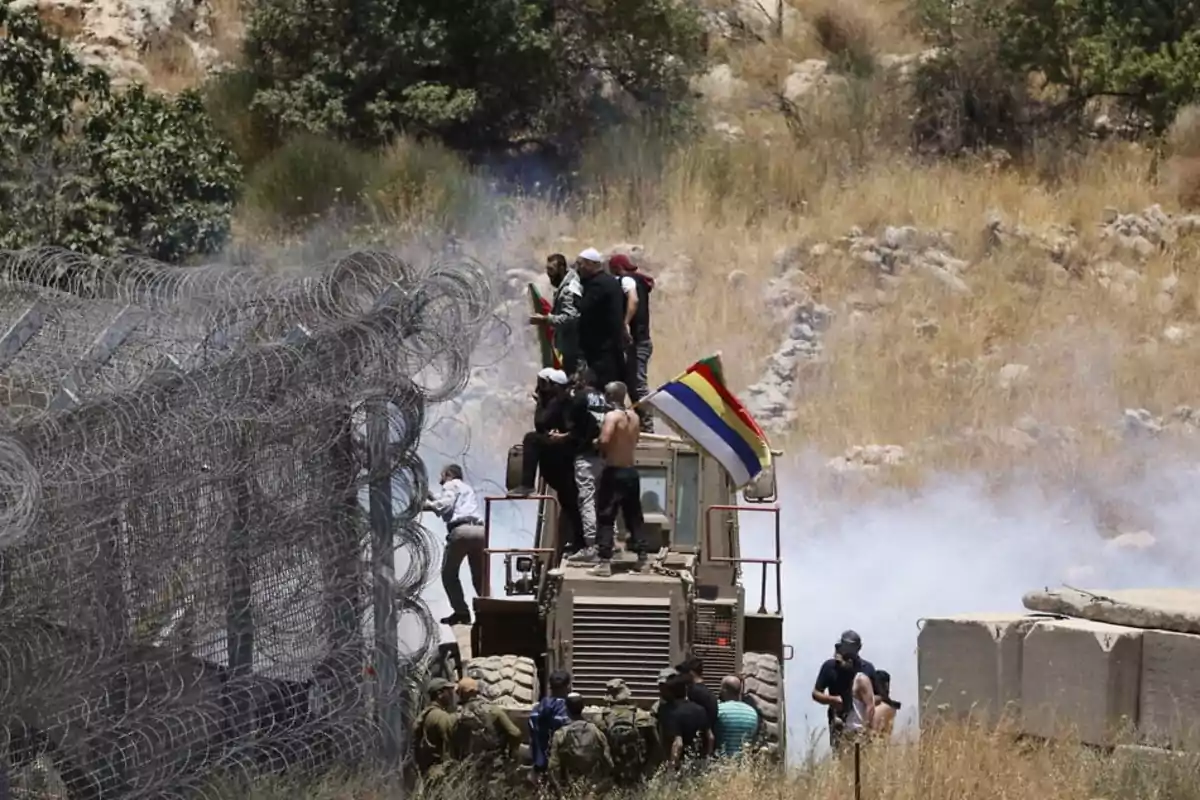 People on an armored vehicle next to a barbed wire fence in an arid area with smoke and a waving multicolored flag