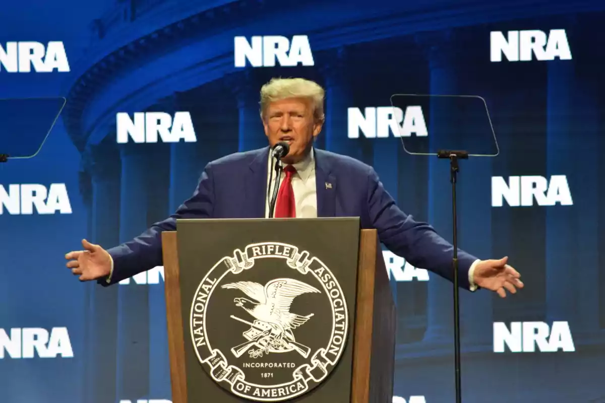 Hombre de cabello rubio con traje azul y corbata roja hablando en un podio de la Asociación Nacional del Rifle con fondo azul y letras NRA Hombre de cabello rubio con traje azul y corbata roja hablando en un podio de la Asociación Nacional del Rifle con fondo azul y letras NRA