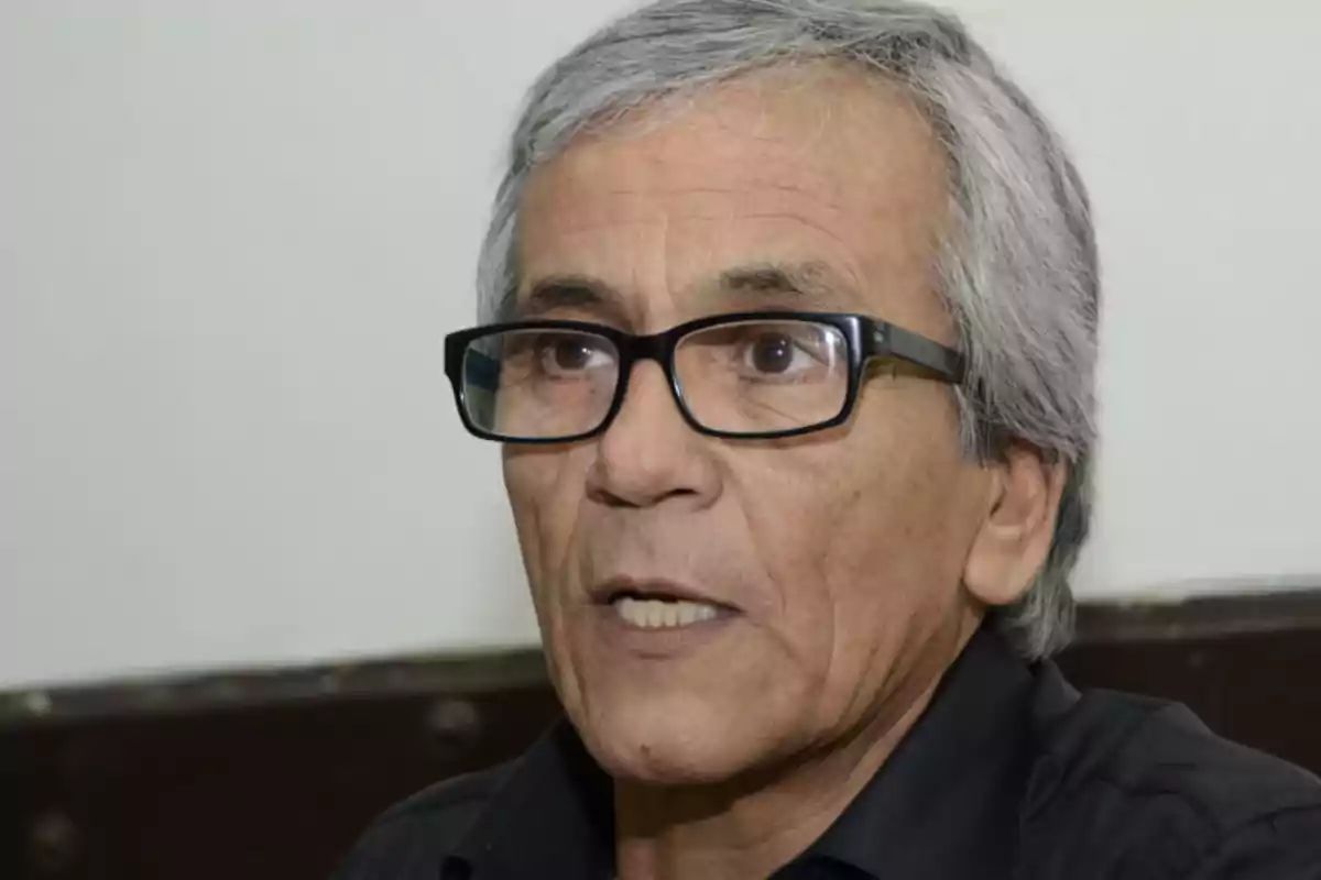 Hombre de cabello canoso y gafas negras hablando, viste camisa oscura y está en un interior con pared clara de fondo
