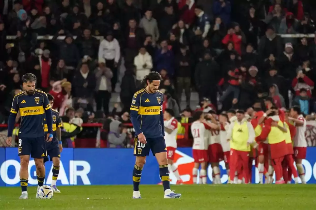 Jugadores de Boca Juniors cabizbajos en el campo mientras el equipo rival celebra al fondo con su afición en las gradas Jugadores de Boca Juniors cabizbajos en el campo mientras el equipo rival celebra al fondo con su afición en las gradas