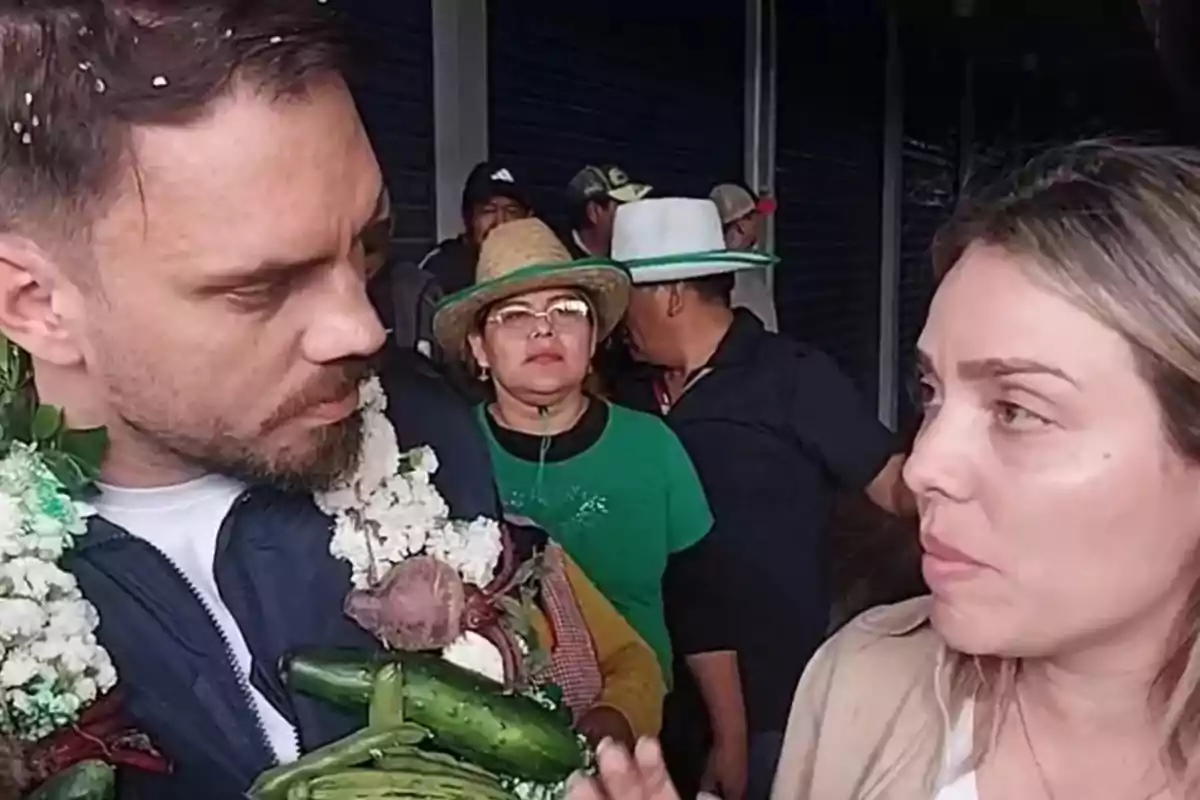 Un hombre con guirnalda de flores y verduras conversa con una mujer mientras varias personas observan al fondo