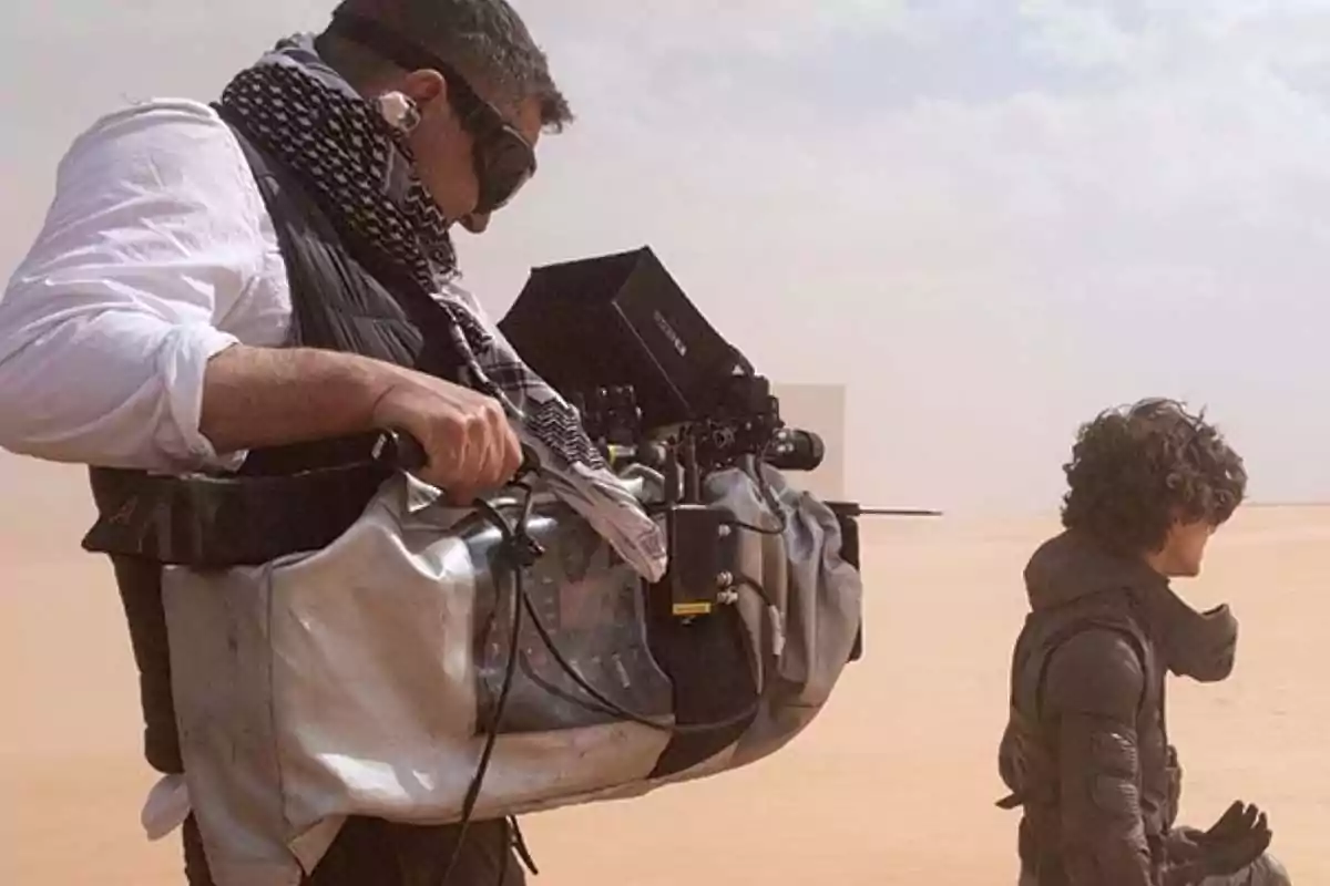 Man with filming equipment and protective goggles in a desert next to a person in dark clothing Man with filming equipment and protective goggles in a desert next to a person in dark clothing