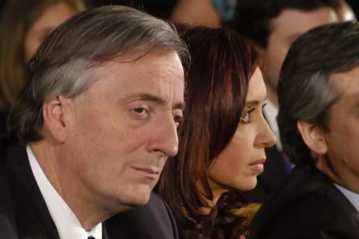 Three people seated in the foreground with serious expressions at a formal event Three people seated in the foreground with serious expressions at a formal event