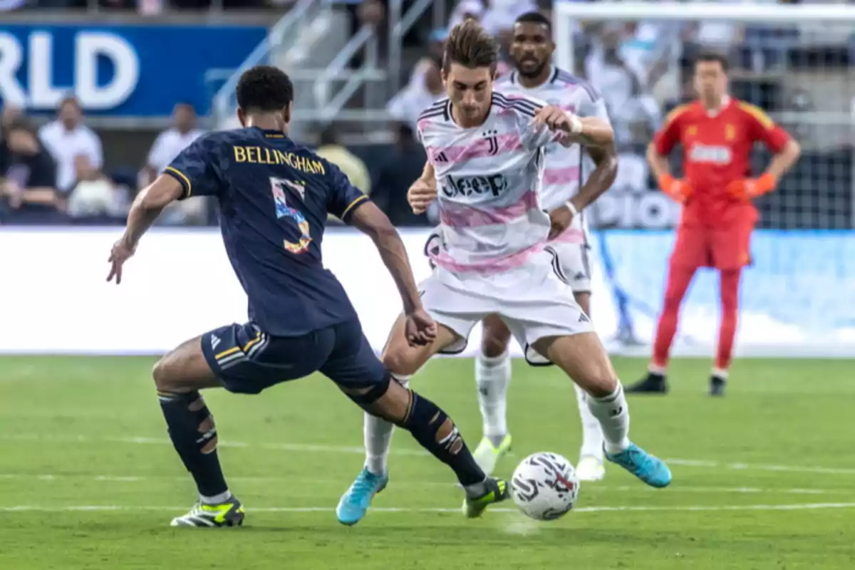 Dos futbolistas disputan el balón durante un partido mientras un portero observa desde el fondo