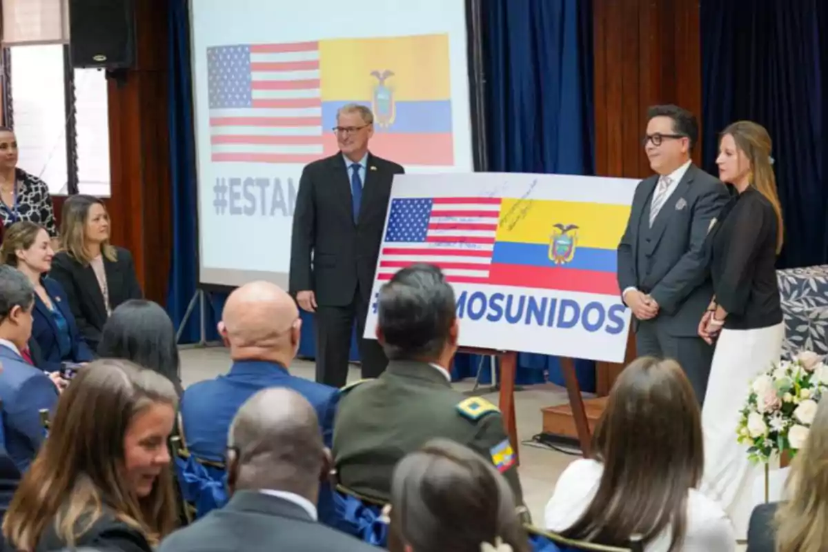 Personas en un evento formal con un cartel que muestra las banderas de Estados Unidos y Ecuador junto al texto estamos unidos mientras varias personas observan sentadas
