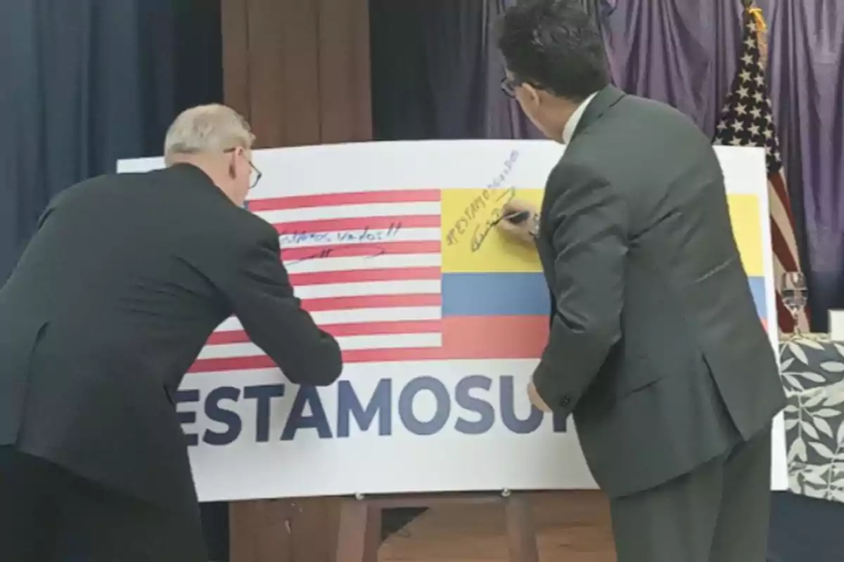 Dos hombres de traje firman un cartel grande que muestra las banderas de Estados Unidos y Colombia junto a la palabra ESTAMOSU Dos hombres de traje firman un cartel grande que muestra las banderas de Estados Unidos y Colombia junto a la palabra ESTAMOSU