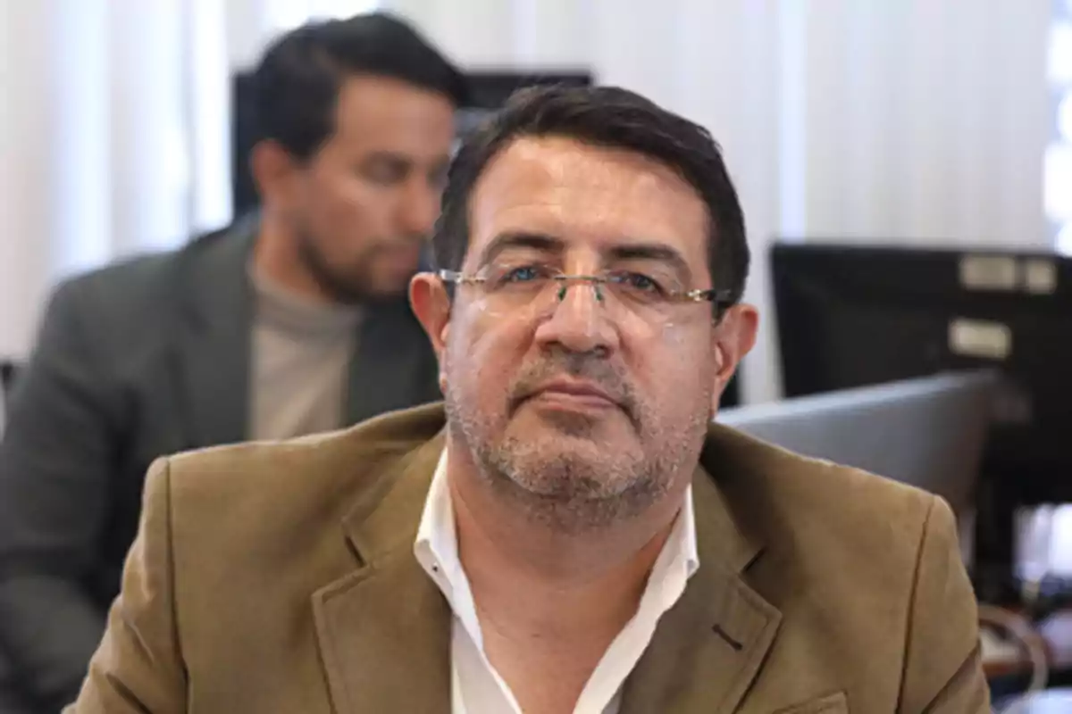 Hombre de mediana edad con gafas y barba corta, vistiendo saco marrón claro y camisa blanca, sentado en una oficina con otra persona desenfocada al fondo Hombre de mediana edad con gafas y barba corta, vistiendo saco marrón claro y camisa blanca, sentado en una oficina con otra persona desenfocada al fondo