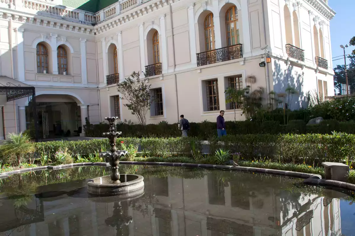 Fuente de agua en el centro de un jardín rodeado de arbustos frente a un edificio antiguo de fachada blanca con ventanas altas y balcones de hierro forjado Fuente de agua en el centro de un jardín rodeado de arbustos frente a un edificio antiguo de fachada blanca con ventanas altas y balcones de hierro forjado
