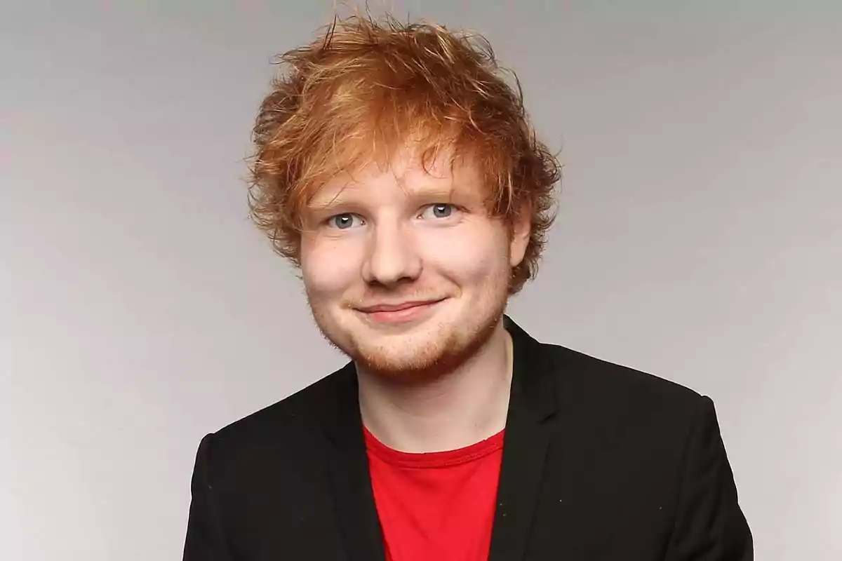 Persona joven de cabello pelirrojo y corto, con barba clara, vistiendo camiseta roja y saco negro, posando frente a un fondo gris claro Persona joven de cabello pelirrojo y corto, con barba clara, vistiendo camiseta roja y saco negro, posando frente a un fondo gris claro