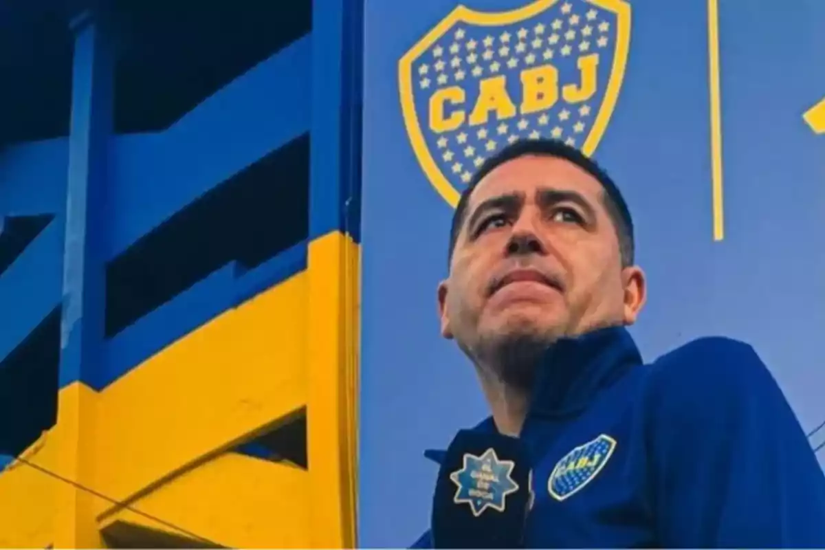 Hombre con chaqueta azul de Boca Juniors frente a una pared azul y amarilla con el escudo de CABJ Hombre con chaqueta azul de Boca Juniors frente a una pared azul y amarilla con el escudo de CABJ