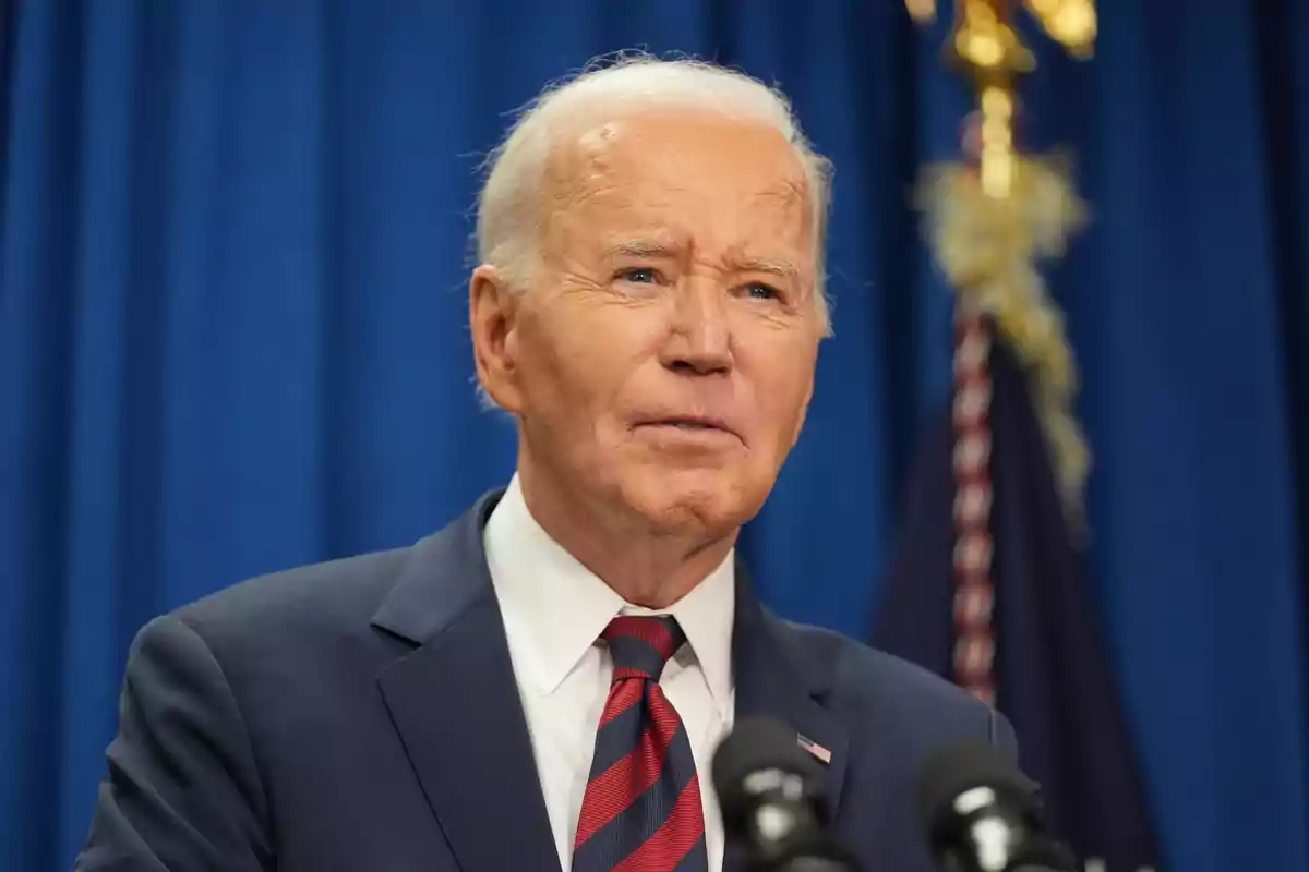 Gray-haired man in a suit and tie speaking in front of microphones with a blue background and a flag behind him Gray-haired man in a suit and tie speaking in front of microphones with a blue background and a flag behind him
