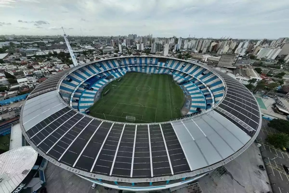 Aerial view of a soccer stadium with light blue and white stands, surrounded by buildings and a city in the background.