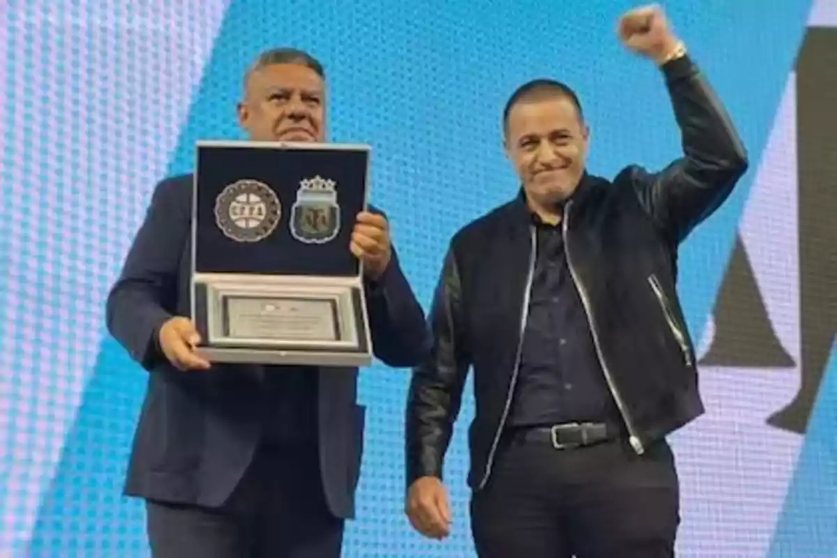 Two men on a stage, one holding a plaque with emblems and the other raising his fist in celebration. Two men on a stage, one holding a plaque with emblems and the other raising his fist in celebration.