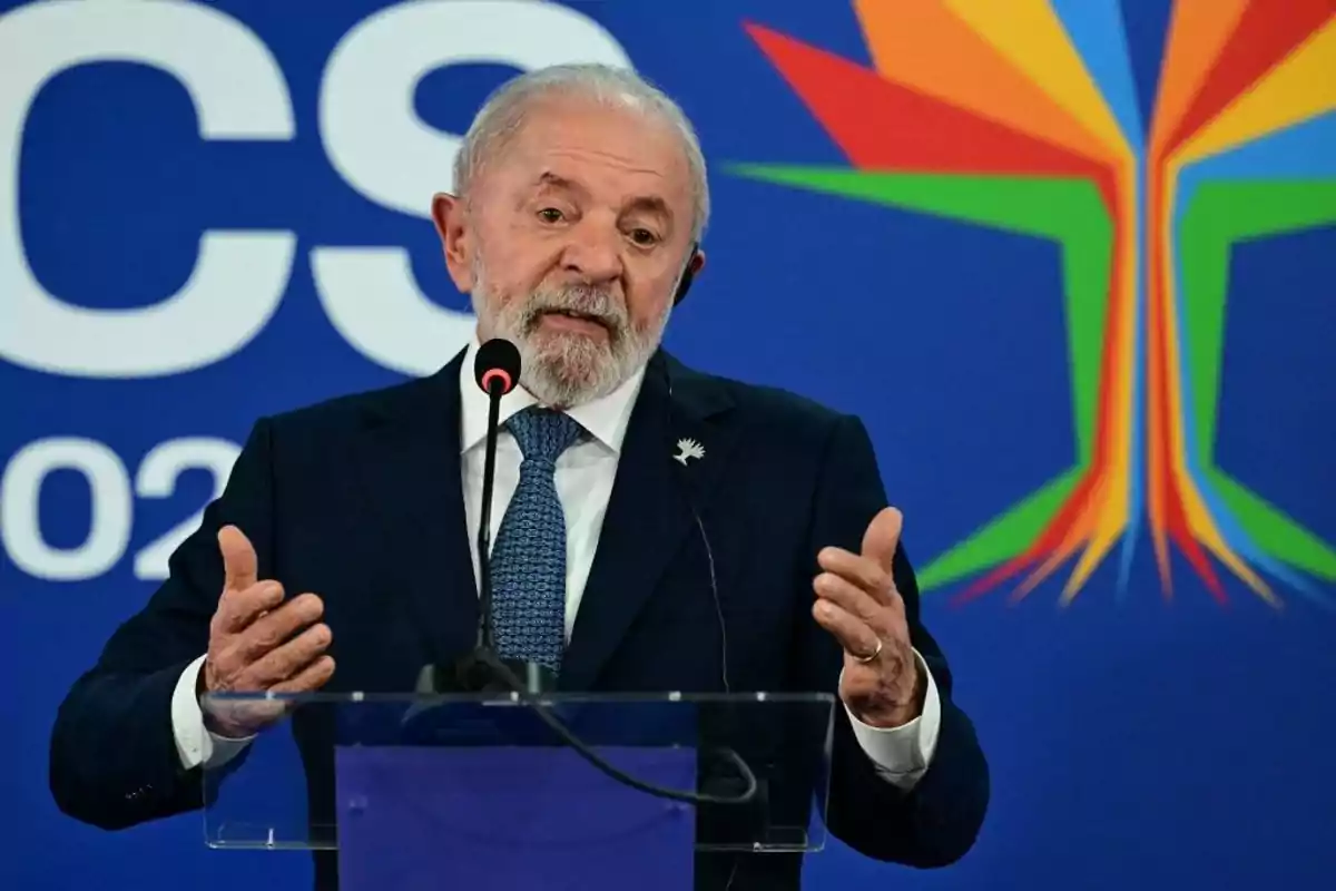 Hombre de cabello canoso y barba hablando en un podio con micrófono frente a un fondo azul con letras blancas y un diseño colorido de líneas geométricas.