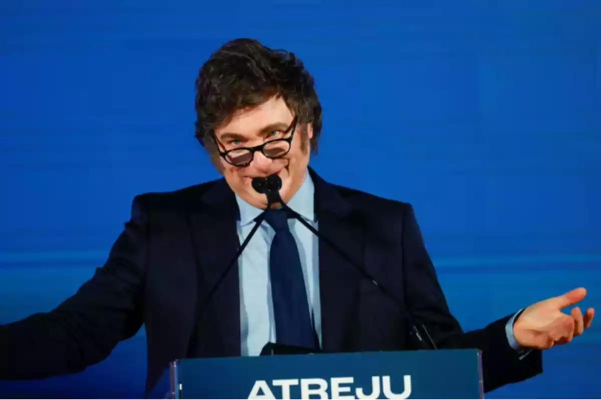 Hombre de cabello oscuro y gafas hablando en un podio azul con fondo azul
