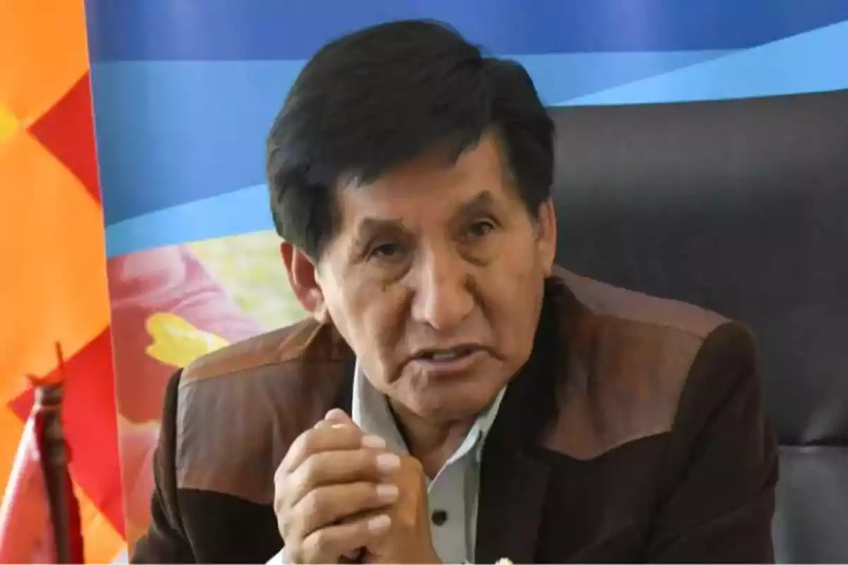 Hombre de cabello oscuro y chaqueta marrón sentado frente a un fondo colorido con las manos entrelazadas Hombre de cabello oscuro y chaqueta marrón sentado frente a un fondo colorido con las manos entrelazadas
