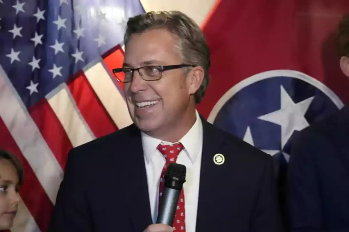 Hombre de traje y corbata roja con micrófono en la mano sonriendo frente a banderas de Estados Unidos y Tennessee Hombre de traje y corbata roja con micrófono en la mano sonriendo frente a banderas de Estados Unidos y Tennessee
