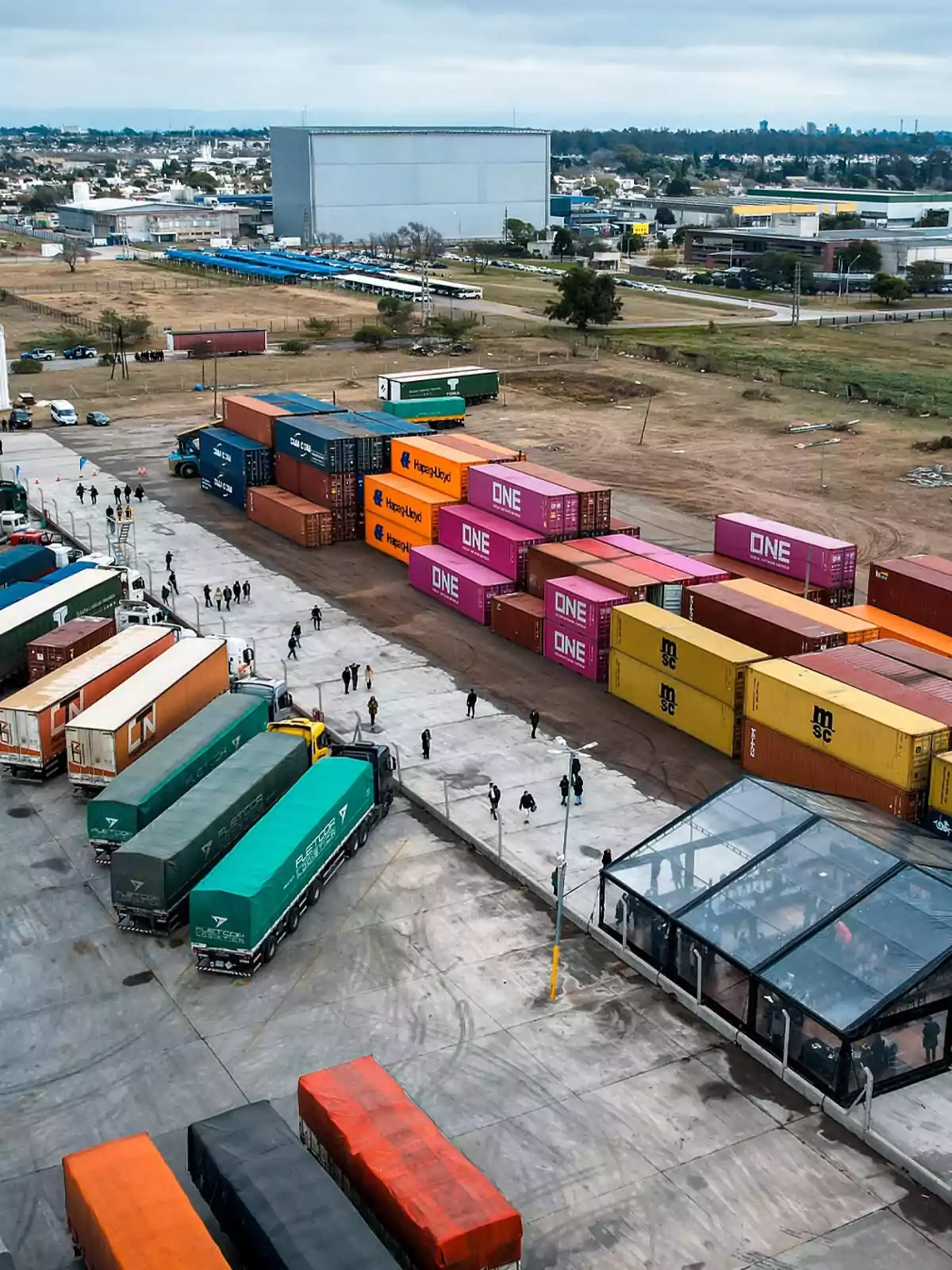 Vista aérea de un depósito logístico con varios camiones y contenedores de colores organizados, personas caminando por el área y edificios industriales al fondo Vista aérea de un depósito logístico con varios camiones y contenedores de colores organizados, personas caminando por el área y edificios industriales al fondo