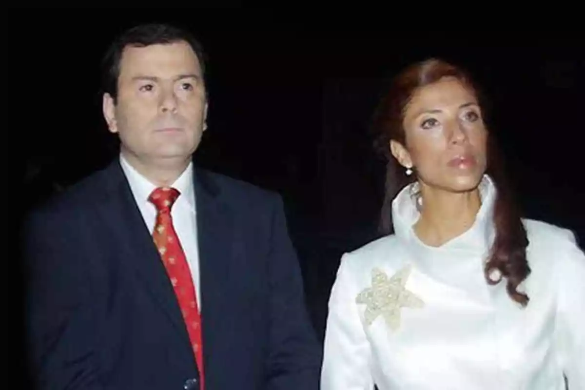 Un hombre con traje oscuro y corbata roja junto a una mujer con vestido blanco y broche en forma de flor sobre un fondo negro Un hombre con traje oscuro y corbata roja junto a una mujer con vestido blanco y broche en forma de flor sobre un fondo negro
