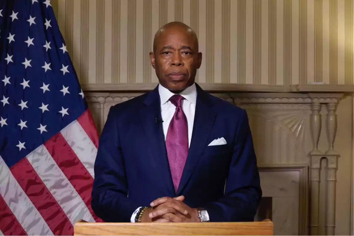 Man in a blue suit and purple tie standing in front of a podium with a United States flag in the background Man in a blue suit and purple tie standing in front of a podium with a United States flag in the background