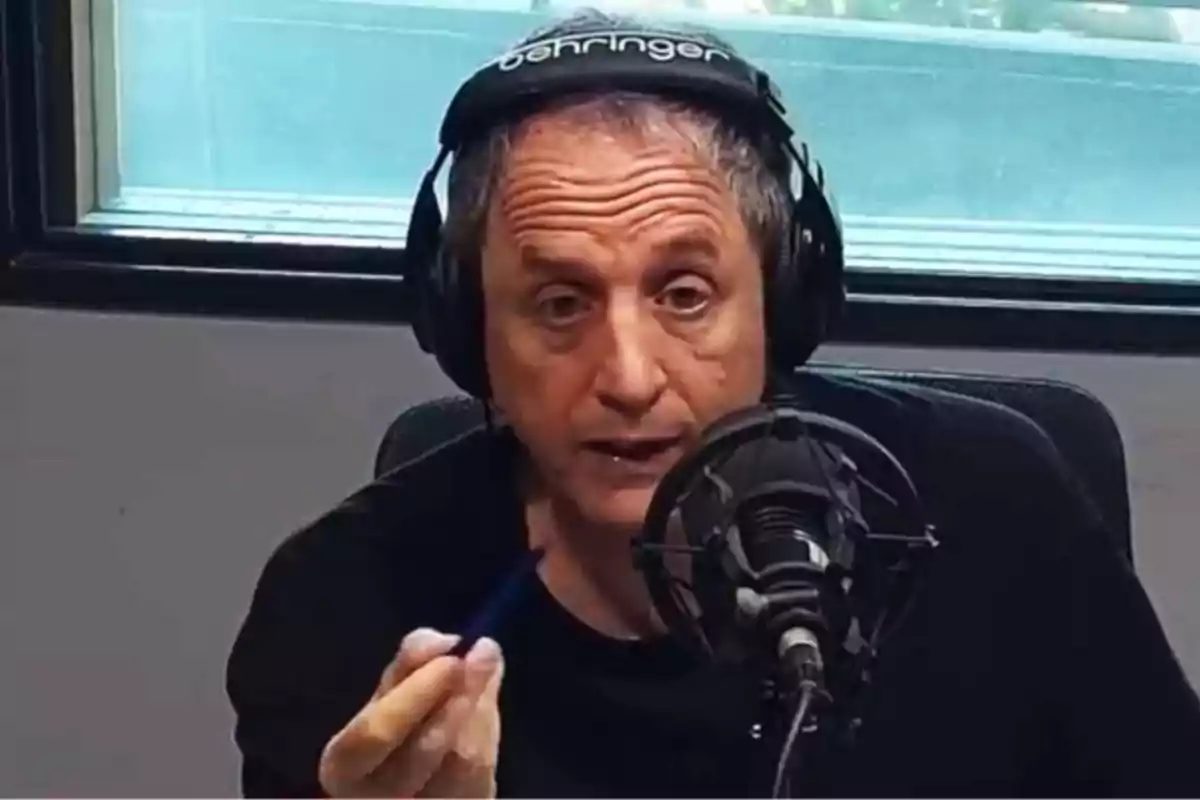 Hombre mayor con audífonos hablando frente a un micrófono en una cabina de radio mientras sostiene un bolígrafo en la mano