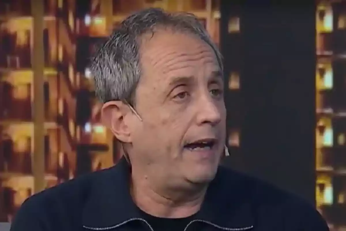 Gray-haired man speaking on a television set with a blurred background of yellow lights