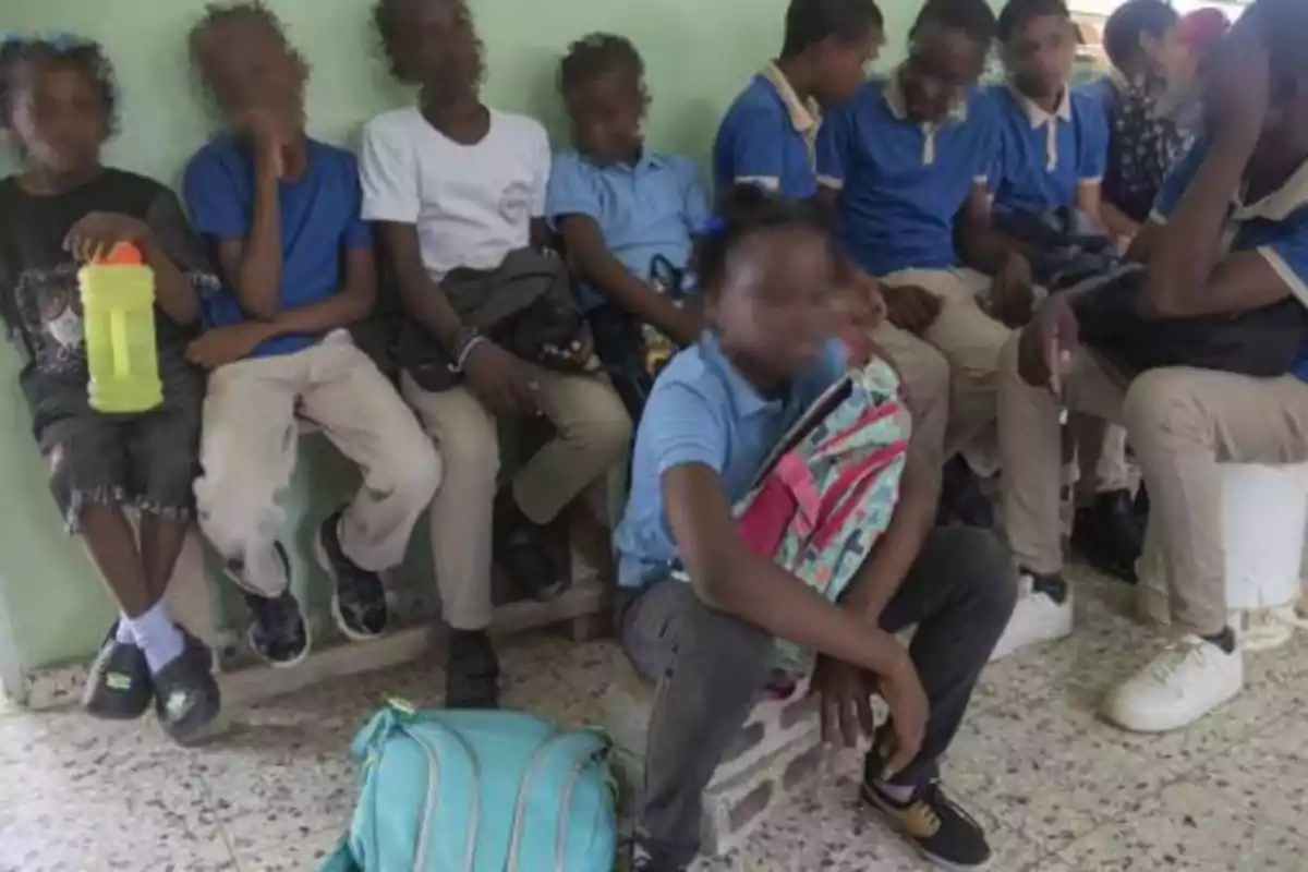 Varios niños con uniformes escolares están sentados en una banca dentro de un aula, algunos con mochilas y botellas de agua, mientras esperan.