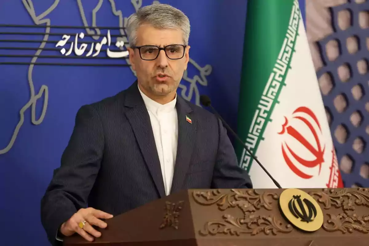 Hombre de cabello canoso y gafas hablando en un podio con el escudo de Irán y una bandera iraní de fondo Hombre de cabello canoso y gafas hablando en un podio con el escudo de Irán y una bandera iraní de fondo
